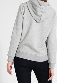 Donna che indossa una felpa cappuccio grigia chiara e jeans scuri, mostrata di spalle con le braccia rilassate lungo i fianchi.