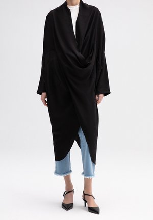 Cardigan noir drapé avec une coupe décontractée, doté de manches longues et d'un ourlet asymétrique. Associé à un jean bleu clair raccourci et des talons noirs.