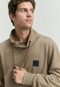 Bruine pullover met een hoge kraag, voorzien van een trekkoord en een donkere vlek op de borst. De stof lijkt zacht met een subtiele textuur.