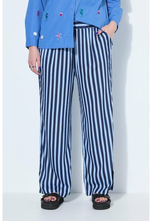 Person trägt blau-weiße vertikal gestreifte, weit geschnittene Hosen, blau besticktes Hemd, schwarze Plateau-Sandalen und lila Nagellack.