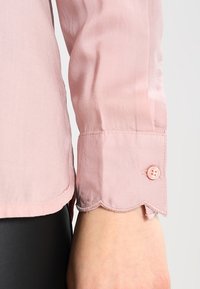 Bluse aus leichtem rosa Seidenstoff mit schimmernden Ärmeln, die einen gewellten Bund und einen einzelnen Knopfverschluss aufweist. Der Stoff zeichnet sich durch eine glatte Textur und dezenten Glanz aus.