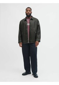 Musta nahkainen vetoketjullinen takki, jossa on suuri kaulus, päällä maroon-paita, yhdistettynä löysiin tummiin housuihin ja mustiin kenkiin, seisoo valkoista taustaa vasten.