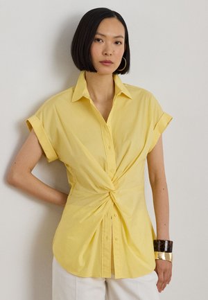 Kobieta o prostych czarnych włosach nosi żółtą koszulę na krótki rękaw zapinaną na guziki, skręconą w talii, białe spodnie oraz złote i ciemne bransoletki.