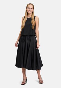 Schwarzes, ärmelloses Top mit subtilen Punktmuster und fließendem schwarzen Rock. Glatte Textur mit lockerer Passform. Getragen mit schwarzen, trägerlosen High Heels.