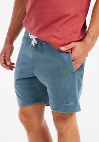 Shorts en coton bleu avec poches latérales, cordon blanc et logo "PRO TEST" en blanc sur l'ourlet. Associé à un T-shirt rouge.