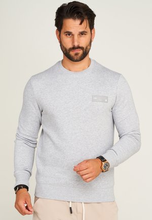 Grauer Pullover mit langen Ärmeln, Rundhalsausschnitt und einem rechteckigen Logo-Patch auf der Brust. Weicher, strukturierter Stoff, lässige Passform.