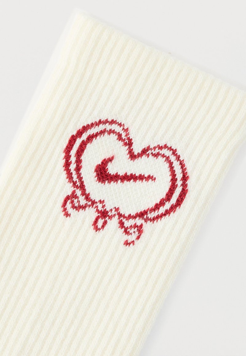 Cream-colored ribbed sock featuring a red embroidered heart design with a swoosh element. Soft texture and elastic material.