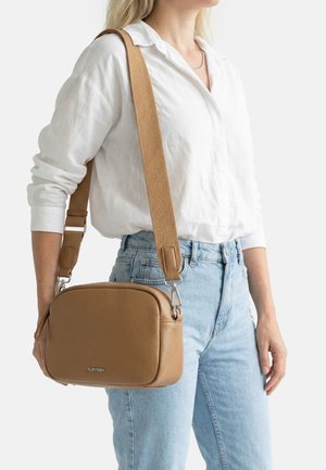 Donna che indossa una camicia bianca e jeans azzurri chiari, con una borsa a tracolla in pelle color cuoio con una fascia larga sulla spalla.