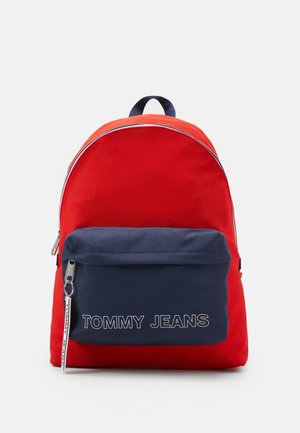 Roter Rucksack mit marineblauer Fronttasche mit "TOMMY JEANS"-Text und Reißverschluss-Zug auf einfachem hellgrauem Hintergrund.