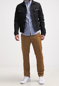 Schwarze Jeansjacke mit zwei Brusttaschen, hellblaues Hemd mit Knöpfen, beige Chinos und weiße Turnschuhe. Fokus auf modernen, lässigen Stil.