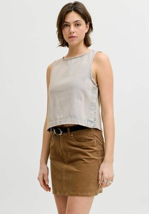 Femme aux cheveux bruns courts portant un haut gris clair sans manches et une jupe en velours côtelé marron avec une ceinture noire, debout devant un fond uni.