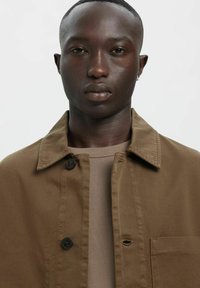 Brązowa kurtka z kołnierzem, zapinana na guziki, z przednią kieszenią, noszona na neutralnej koszuli; wykonana z teksturowanej tkaniny z delikatnym postrzępieniem.