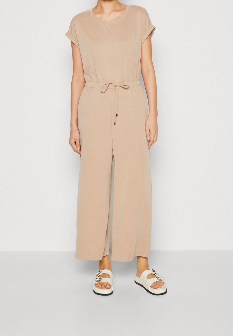 Beige jumpsuit met korte mouwen en een taille met trekkoord; ontwerp met brede pijpen. Draagt witte sandalen met bloemmotieven.