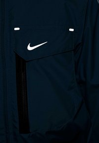 Rompevientos azul marino con un gran bolsillo frontal con cremallera negra, costuras de acento en blanco y un prominente logotipo de Nike en blanco.