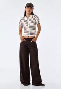 Camiseta polo de manga corta a rayas con cuello en crema, marrón y burdeos; combinada con pantalones de pierna ancha de color marrón oscuro; accessorios incluidos: una gorra marrón.