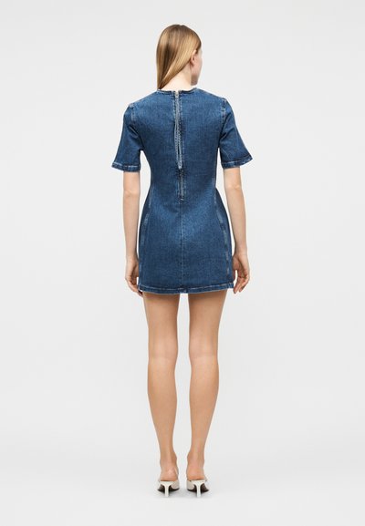 Γυναίκα που στέκεται με την πλάτη, φοράει κοντομάνικο, κοντό τζιν φόρεμα με φερμουάρ στην πλάτη και λευκές ψηλοτάκουνες γόβες.