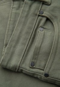 Tissu vert olive avec une texture côtelée visible, comportant une poche à rabat fermée par deux boutons en métal noir et des bords surpiqués.