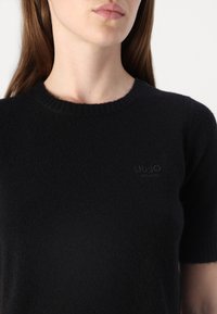 Svart strikket genser med korte ermer, rund hals og ribbestrikket kant. Har en subtil brodert logo på brystet. Myk tekstur.