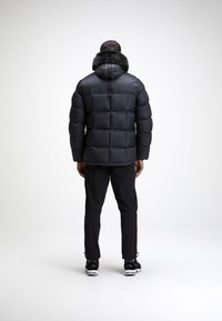 Svart pufferjakke med hette foret med pels, quiltet design og elastiske mansjetter, kombinert med svarte bukser og joggesko.