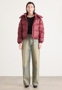 Giacca imbottita bordeaux con colletto alto e polsini a costine, abbinata a pantaloni in denim chiaro a gamba larga. Calzature nere eleganti completano l'outfit.