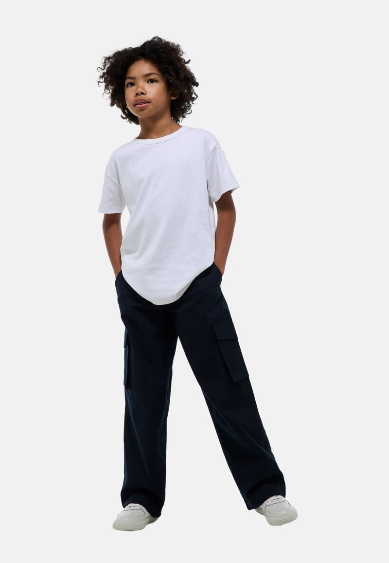 Un enfant avec des cheveux bouclés se tient debout portant un t-shirt blanc uni, un pantalon cargo noir et des baskets blanches, les mains dans les poches, sur un fond clair.