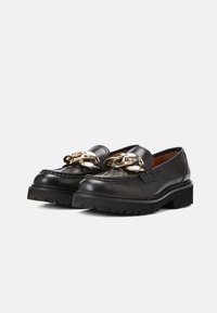 Mocassins en cuir noir avec semelles épaisses et large chaîne dorée sur le devant, présentés côte à côte sur un fond blanc.