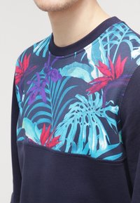 Uomo che indossa una felpa blu navy con un vivace motivo floreale tropicale in blu, rosso e viola sul petto e sulle spalle.