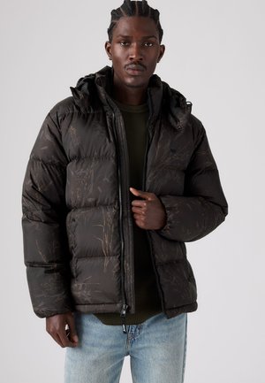 Homme aux cheveux tressés portant une doudoune sombre à motifs par-dessus un pull vert et un jean bleu clair sur un fond uni.