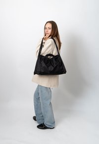 Mujer con chaqueta beige y vaqueros azul claro lleva un gran bolso negro al hombro, de pie frente a un fondo blanco liso.