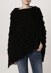 Svart stickad poncho med ett texturerat vågmönster, med lager av volanger. Lös passform, öppna sidor, bärs över ett par blå jeans.