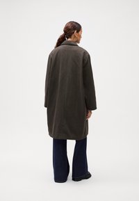 Woman with dark hair in a low ponytail wearing a dark brown long coat, dark blue wide-leg jeans, black shoes, and large hoop earrings, facing away.