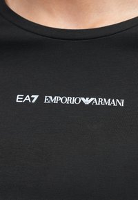 Svart T-skjorte i bomull med rund hals, med en hvit logo som inkluderer "EA7" og "EMPORIO ARMANI" i en fet, moderne font.