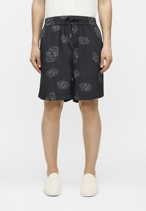 Mężczyzna ubrany w czarne szorty z troczkami z szarymi kwiatowymi wzorami, białe buty wsuwane i białą koszulę, stojący na jednolitym tle.