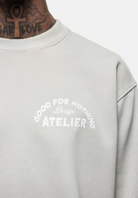 Lichtgrijze sweatshirt met ronde halslijn, voorzien van witte tekst: "GOOD FOR NOTHING Design ATELIER" in een gebogen ontwerp. Zachte textuur.