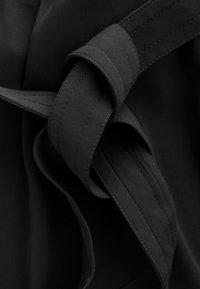 Schwarzer Stoff mit glatter Textur und einer geknoteten Detail. Die Webart ist eng, und die Stiche sind entlang der Kanten des Bandes sichtbar.