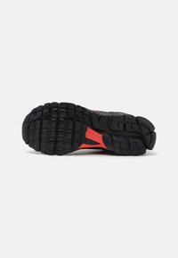 Sport shoe sole with deep treads, predominantly black with red accents. Textured rubber providing grip and stability. Lightweight design.