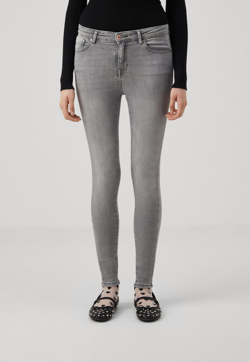 Tajta gråjeans med en slät textur, fem fickor och knappstängning, kombinerat med svarta platta skor och mönstrade strumpor.