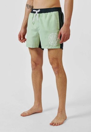 Un short de bain vert et marine avec une taille élastique, un cordon blanc et un logo circulaire sur le devant. Tissu lisse, coupe au-dessus du genou.
