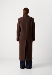 Cappotto lungo in lana marrone con colletto, ventrale posteriore e due tasche laterali. Presenta dettagli con bottoni sulle maniche. Adatto per il clima più fresco.