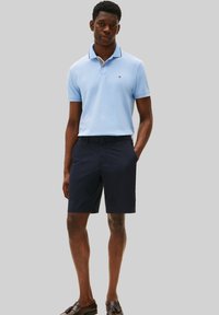 Homme portant un polo bleu clair, un short navy et des mocassins marron, se tenant avec une main dans la poche contre un fond gris uni.