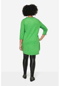 Grüne Tunika mit Dreiviertelärmeln, strukturiertem Stoff und gewelltem Saum, kombiniert mit schwarzen Leggings und mit Nieten besetzten schwarzen Schuhen.