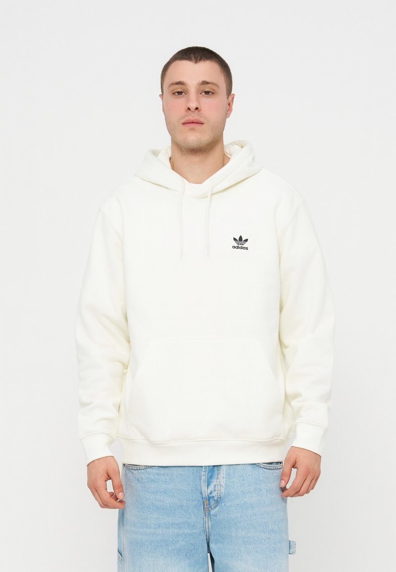 Młody mężczyzna w białej bluzie z kapturem Adidas i jasnoniebieskich dżinsach stoi na gładkim białym tle.
