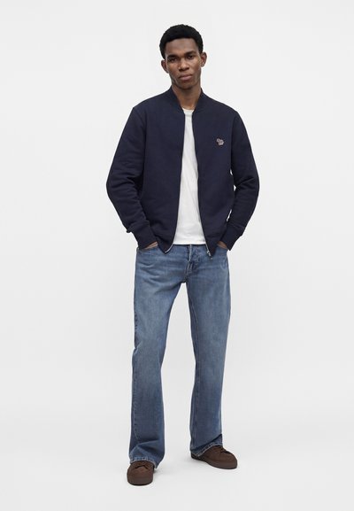 Jeune homme debout, les mains dans les poches, portant une veste bleu marine, un t-shirt blanc, un jean bleu et des chaussures marron sur un fond uni.