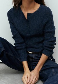 Haut en maille bleu marine à manches longues avec un col arrondi et une patte de boutonnage à trois boutons, associé à un jean en denim foncé. Tissu doux et texturé.