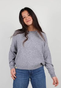 Pull en tricot gris avec un décolleté large et une coupe ample. Poignets et ourlet côtelés. Associé à un jean bleu.