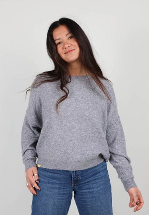 Pull en tricot gris avec un décolleté large et une coupe ample. Poignets et ourlet côtelés. Associé à un jean bleu.