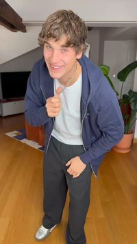 Outfit casual con una felpa blu con zip, T-shirt bianca, pantaloni neri e sneakers argento. Ambiente interno con piante e pavimento in legno.