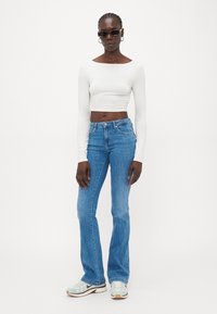 Vit långärmad crop top, ljusblå utsvängda jeans och grå sneakers med gröna detaljer. Modell som bär svarta solglasögon och hoopörhängen.
