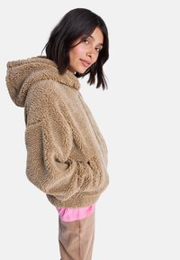 Beige, fluffy hoodie met een pluche textuur, oversized vorm en trekkoordcapuchon. Draag draag over een roze shirt, zijaanzicht tegen een witte achtergrond.