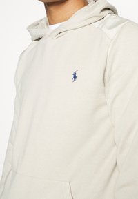 Sweat à capuche beige clair avec un tissu texturé, doté d'une poche kangourou et d'un petit logo bleu sur la poitrine. Capuche avec cordons inclus.